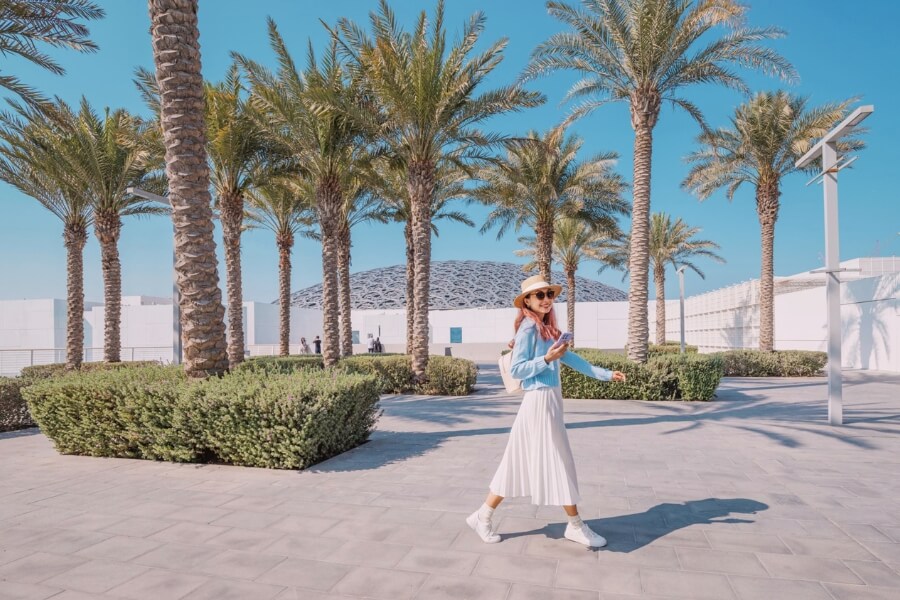 woman walking with her phone in front of Louvre Abu Dhabi in long dress and sun hat culturally respectful clothing for venues in UAE